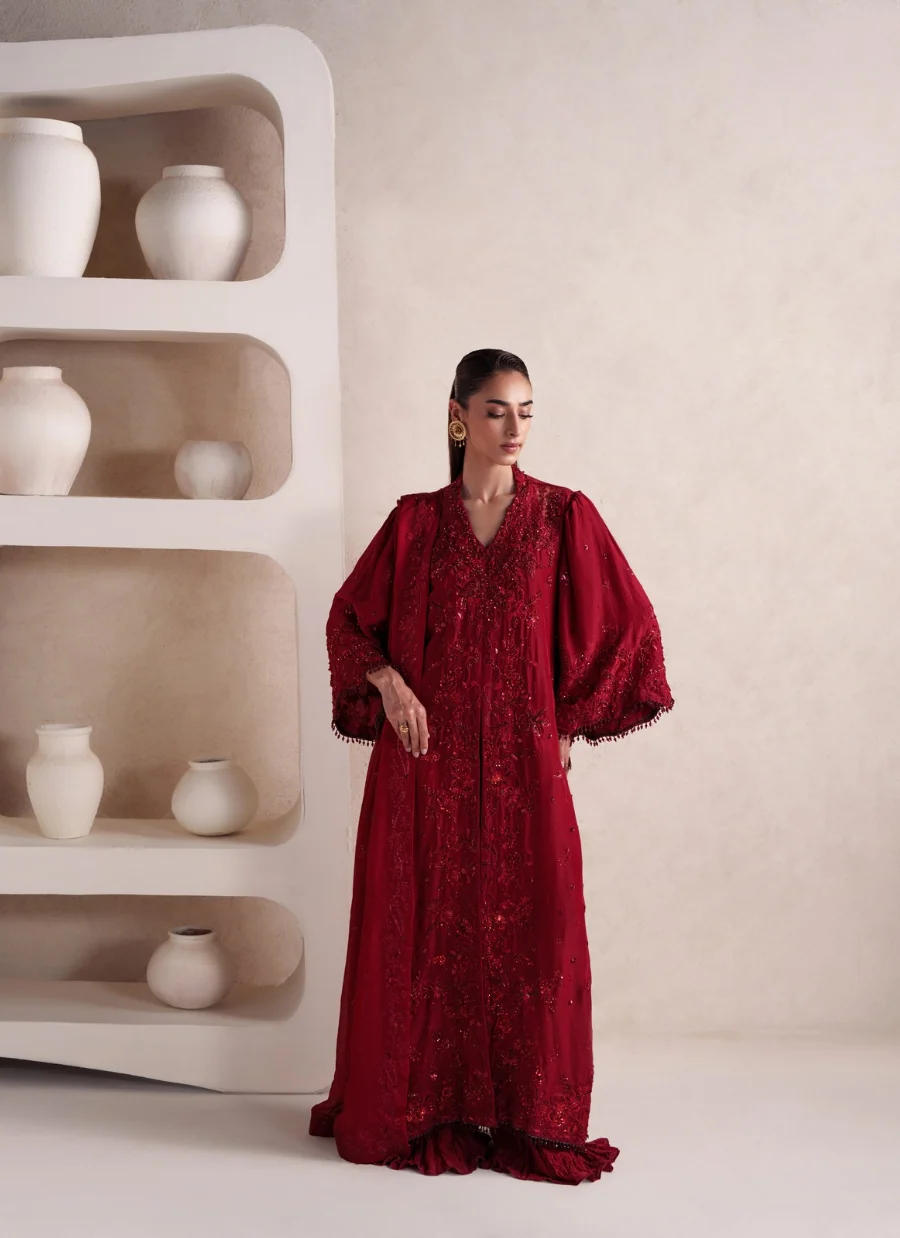 A woman wearing a long, deep red embroidered gown with voluminous sleeves stands in a minimalist studio setting next to a sculptural white shelf filled with white ceramic vases.