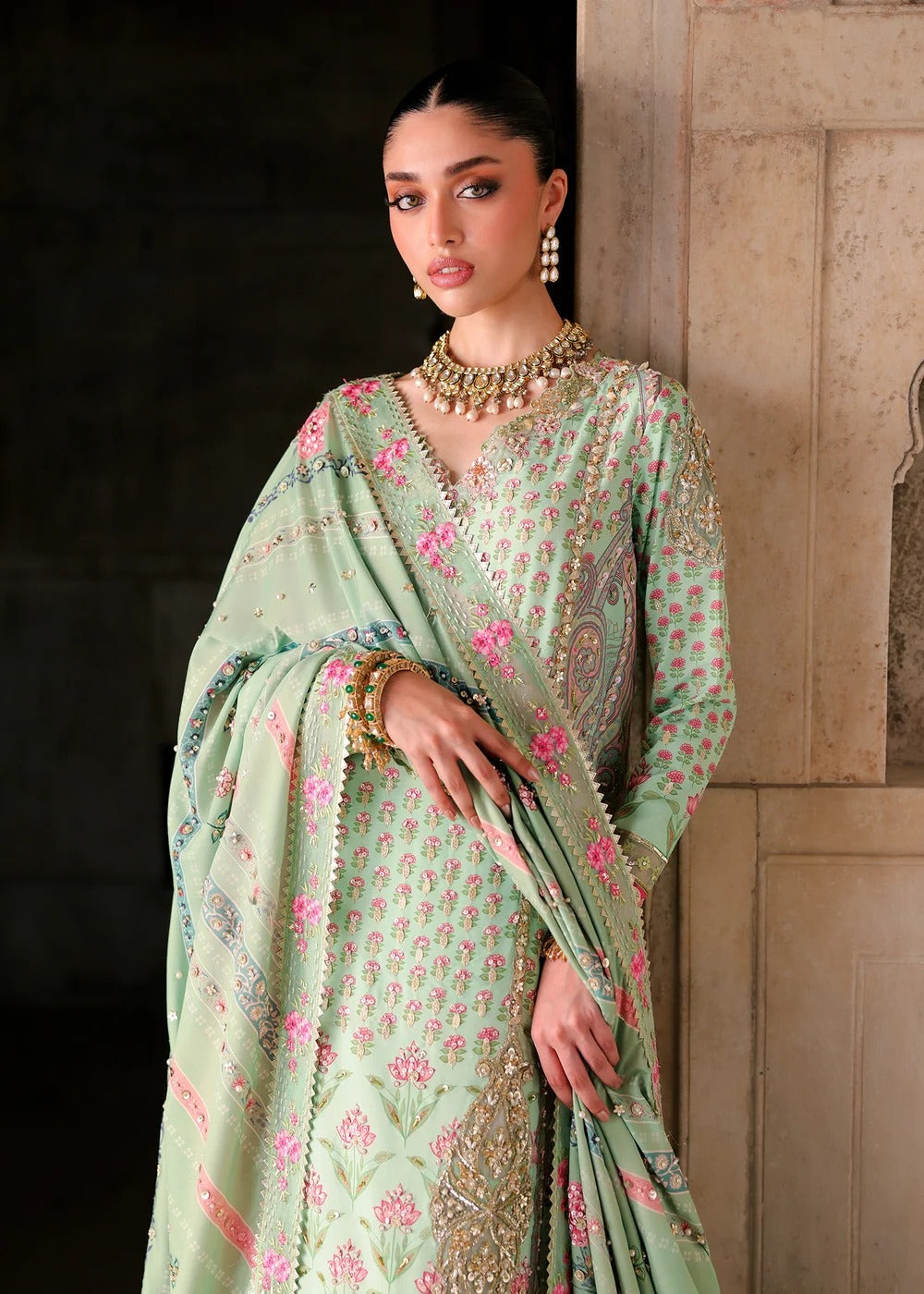 A woman models a mint green embroidered traditional outfit with a matching dupatta, featuring pink floral patterns and gold detailing, set against a dark and light stone wall background.