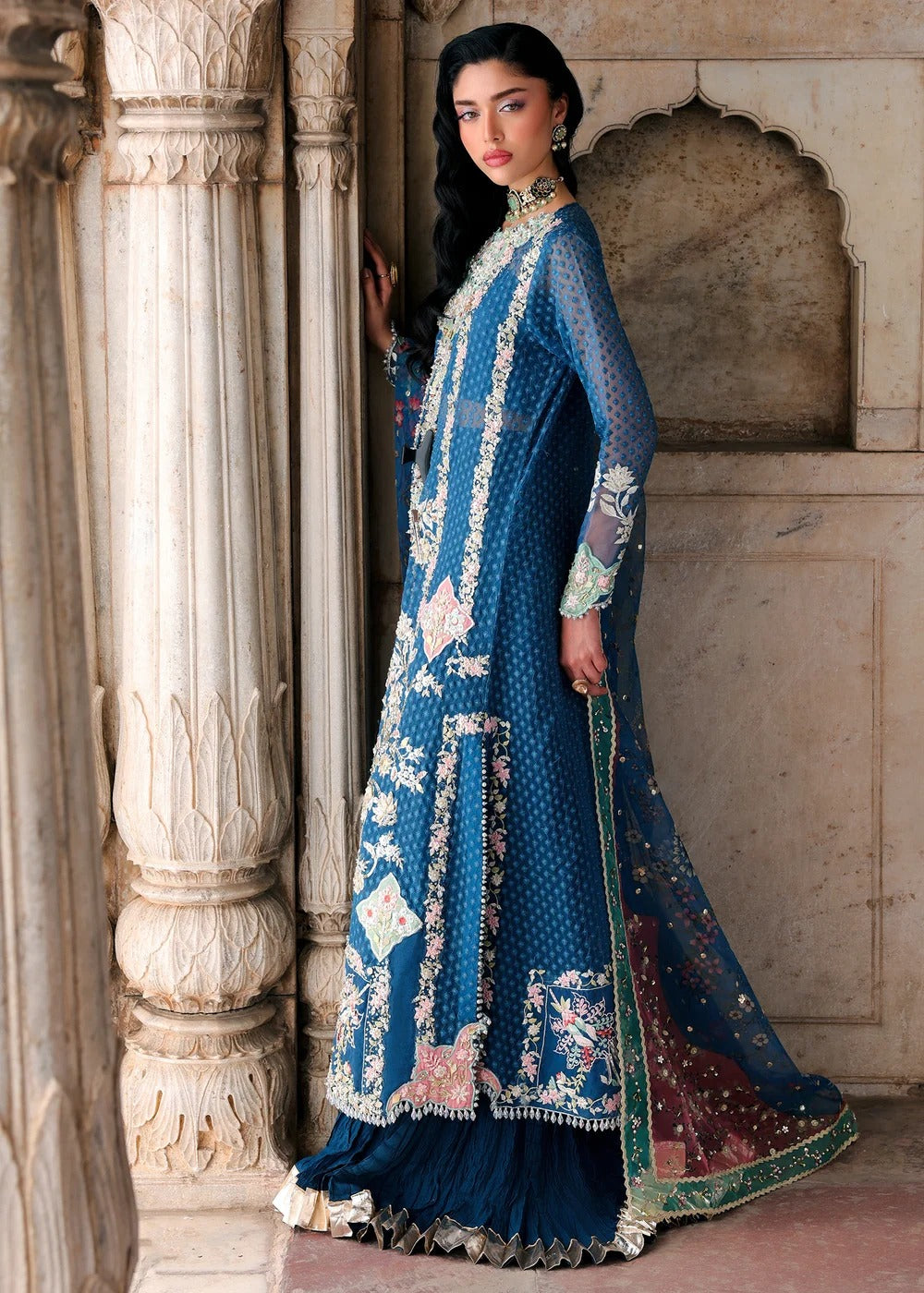 A woman models a traditional blue embroidered Anarkali suit with matching dupatta in an ornate stone architectural setting.