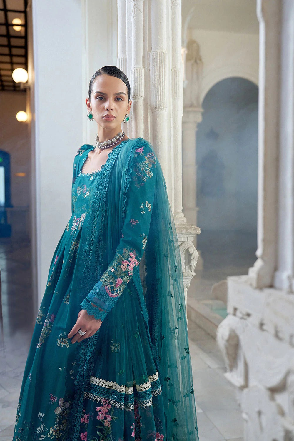 Woman in a teal embroidered saree standing in an elegant indoor setting.