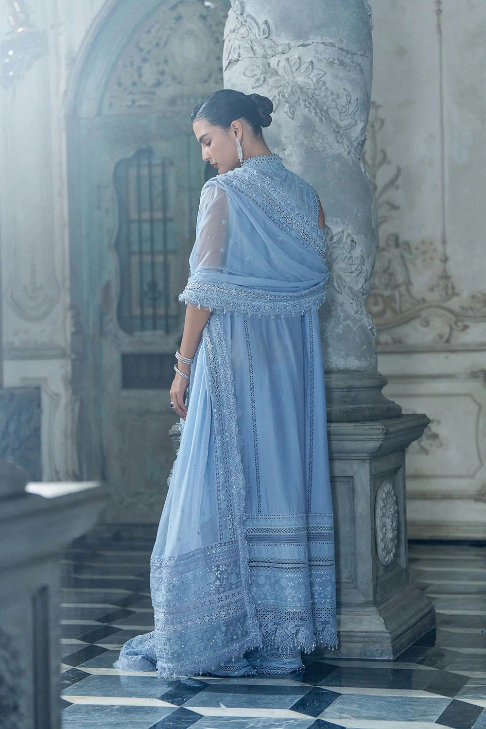 Woman in a light blue traditional outfit standing in an ornate interior setting.