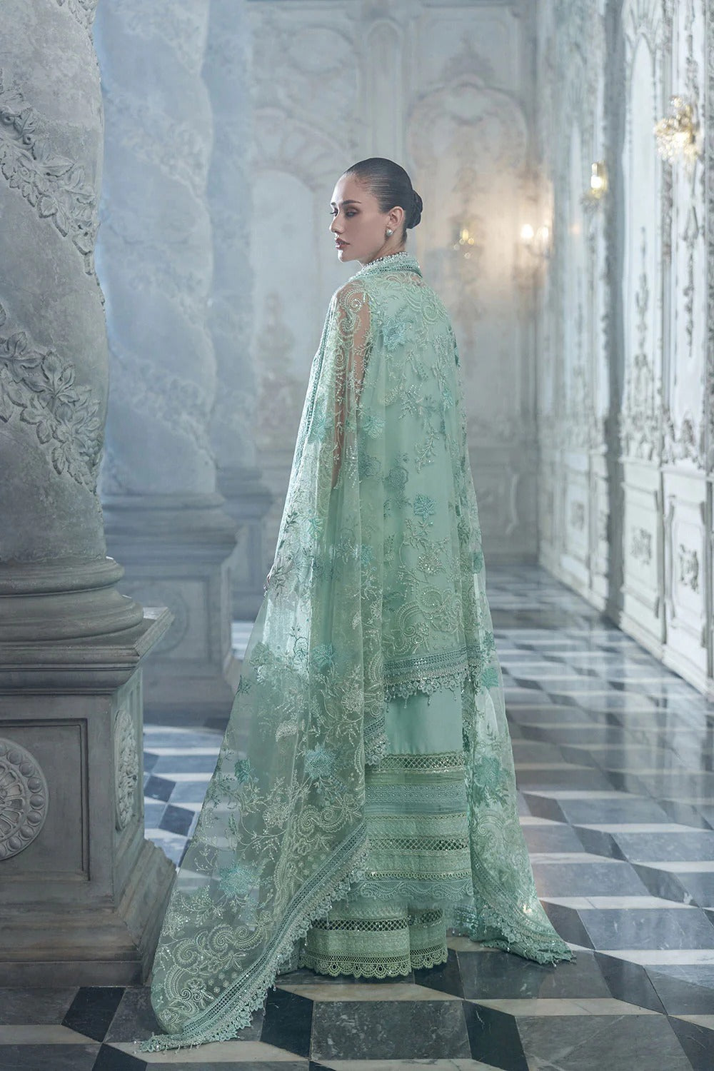 Woman in a light green embroidered traditional outfit standing in an elegant interior setting.