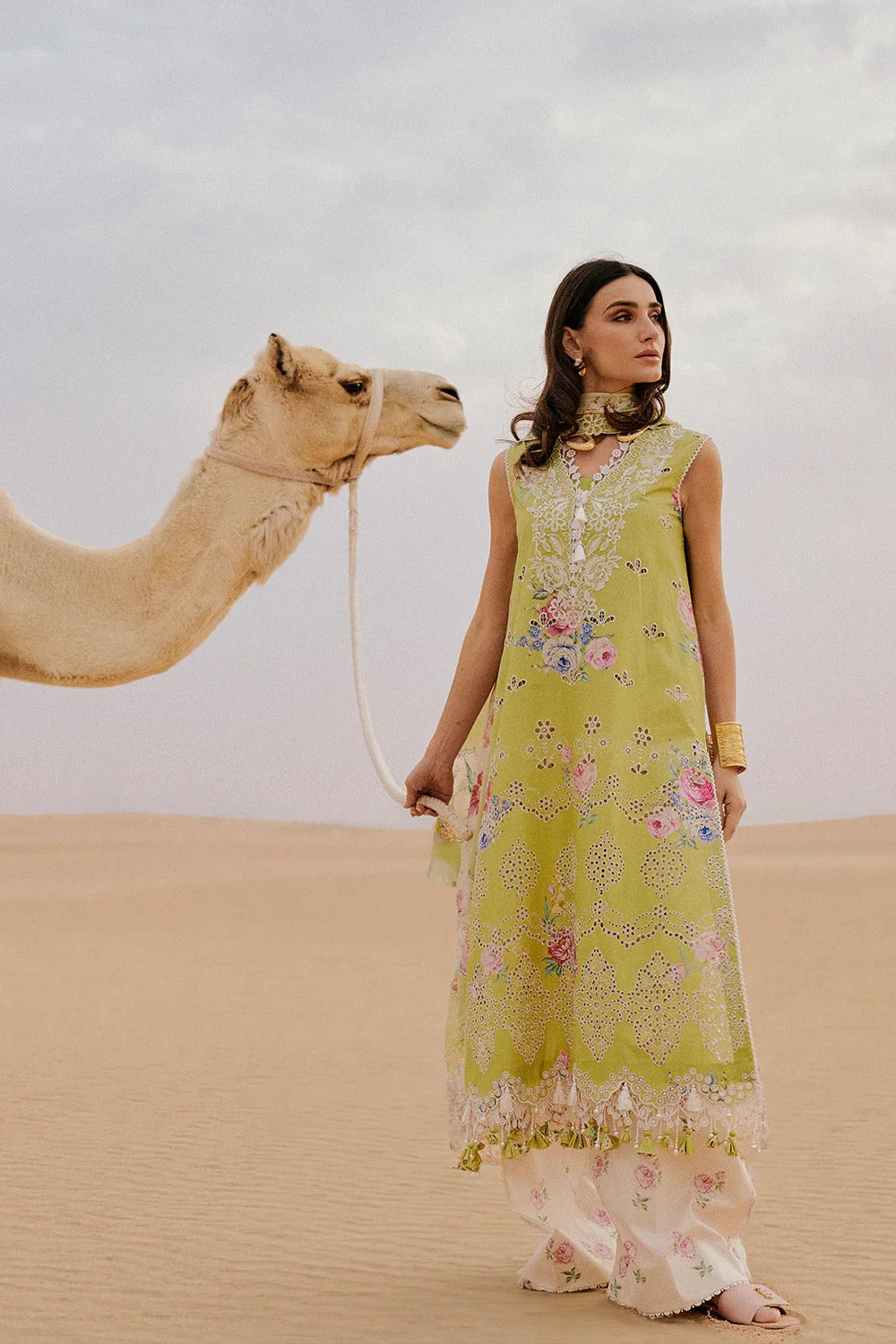 Woman in a green embroidered dress standing next to a camel in a desert setting