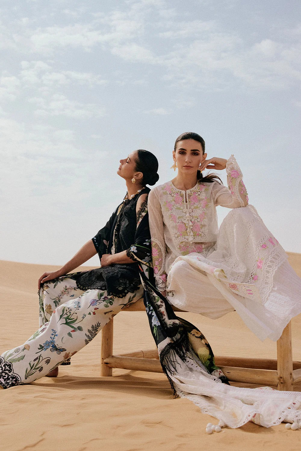 Two women in traditional outfits sitting on a sand dune with a clear sky.