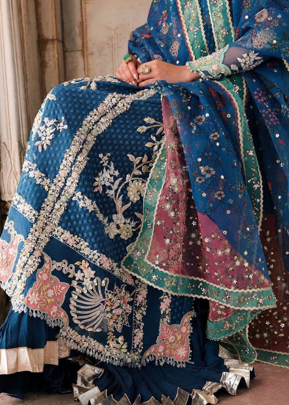 A close-up of a person wearing an ornate traditional outfit with a deep blue embroidered skirt, matching dupatta, and a pink and teal border drape. The fabric features intricate silver and white floral embroidery, sequins, and metallic detailing.