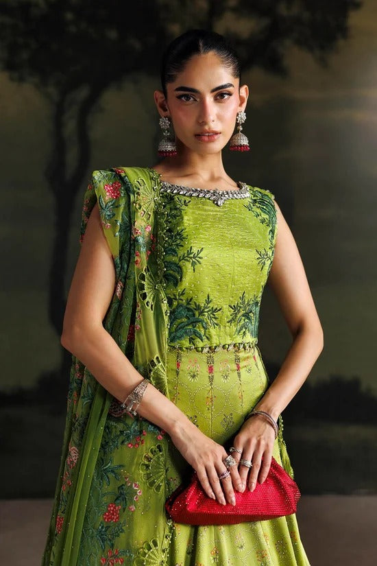 A woman modeling a green embroidered traditional outfit with floral patterns, holding a red clutch purse against a dark, blurred outdoor background.