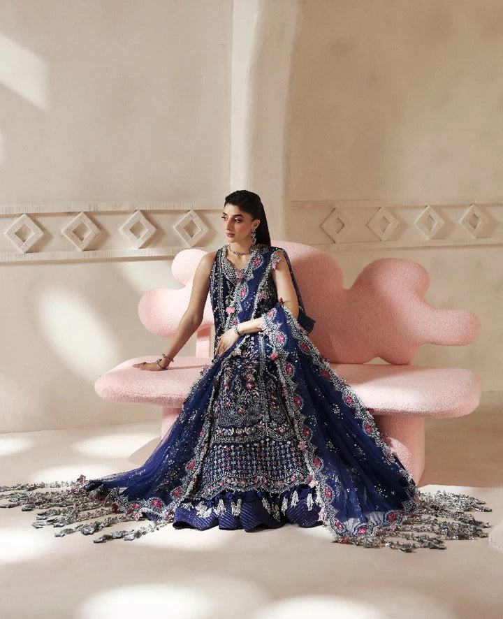 Woman in a blue embroidered traditional outfit sitting on a pink chair with decorative wall behind