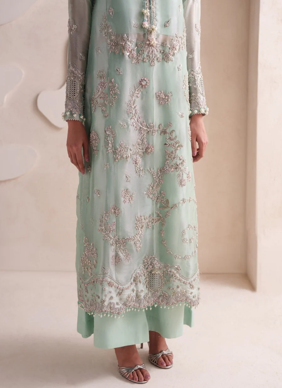 A model wearing a mint green traditional embroidered dress with matching pants and silver sandals, shown from the neck down in a minimalist beige room.