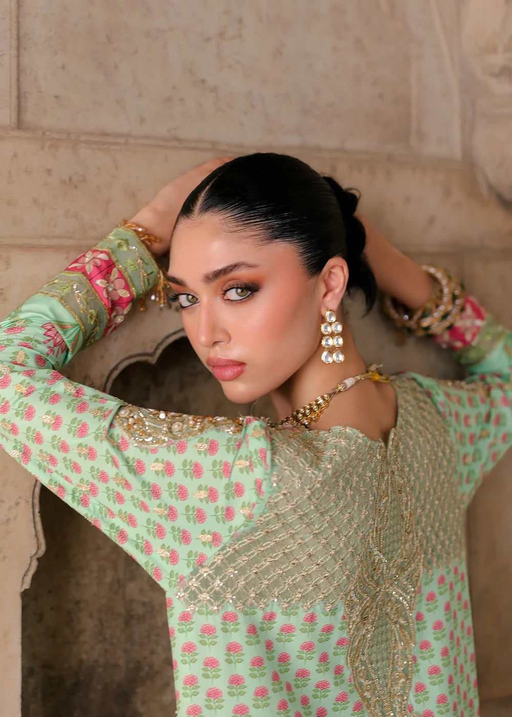 A woman wearing a mint green printed traditional outfit with gold embroidery, posing in front of a stone wall.