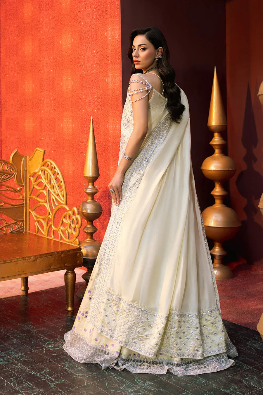 Woman in a white saree with gold details against a red wall with decorative elements.