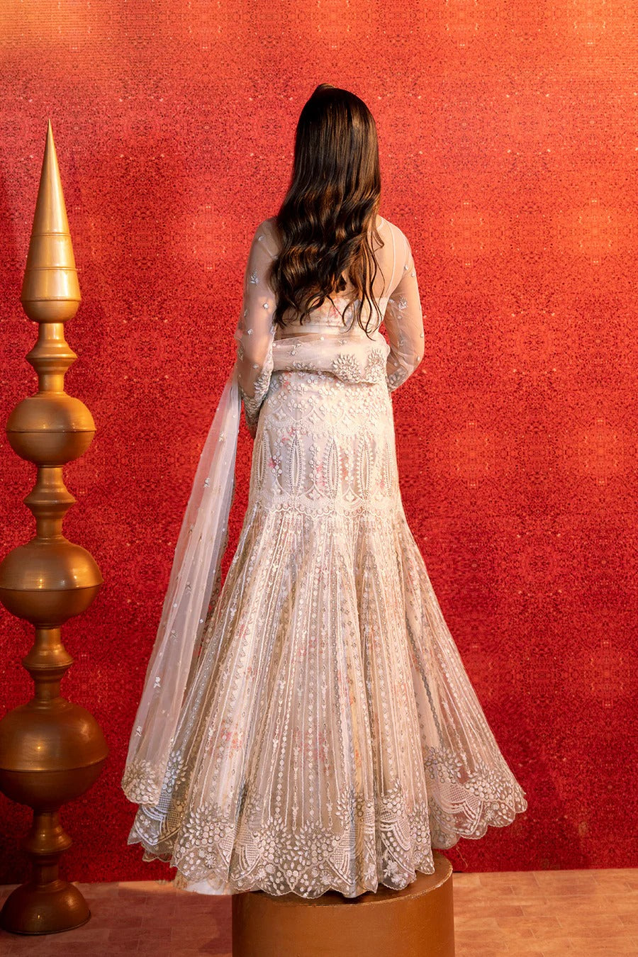 Woman in a white embroidered dress standing against a red wall with a gold decorative item.
