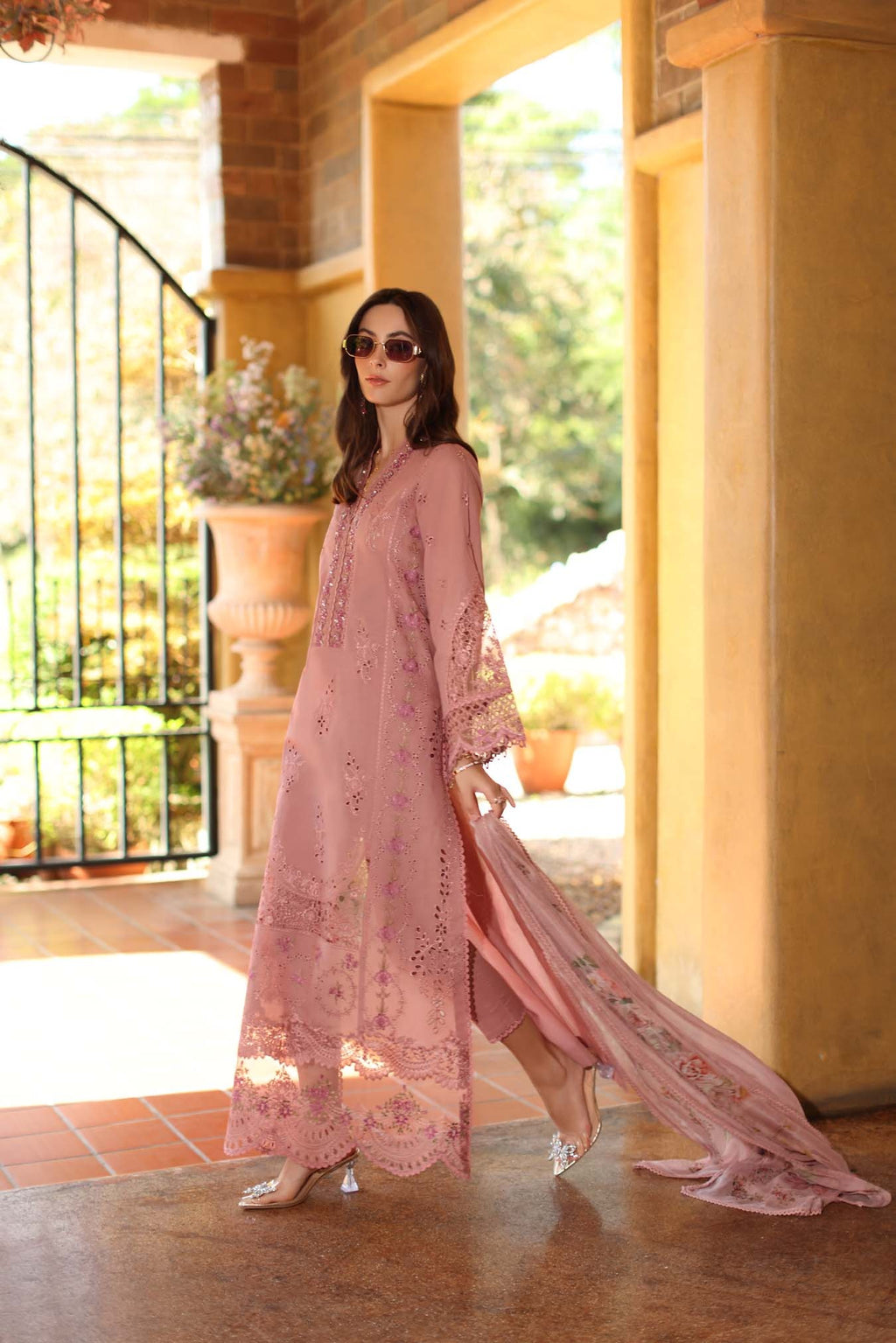 Woman in a pink lace dress standing in a sunlit room with large windows.