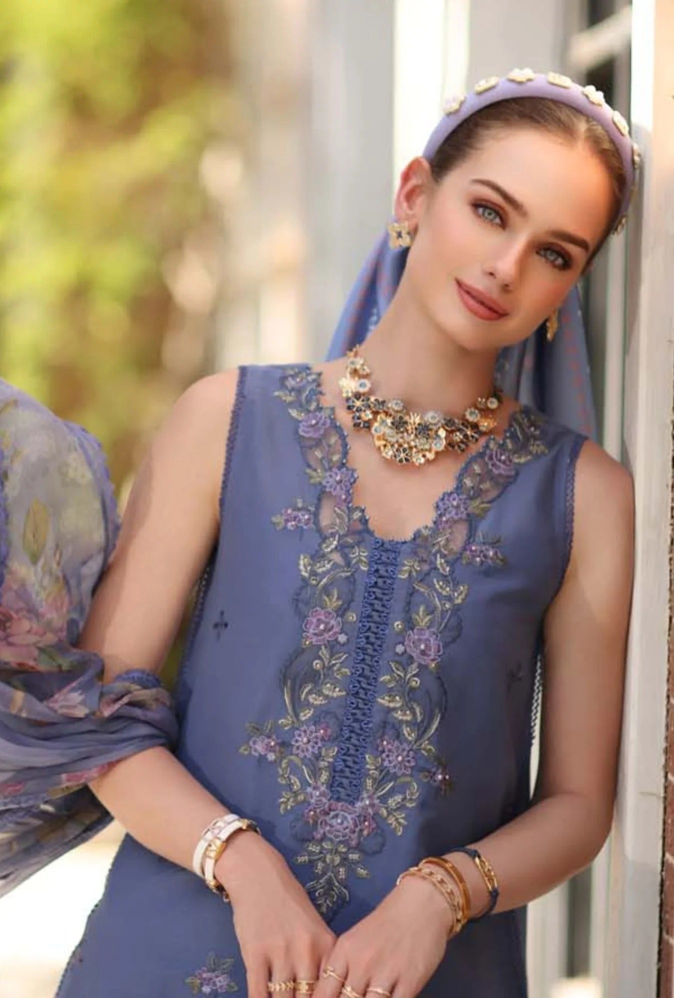 Woman wearing a blue embroidered dress with a matching headscarf, standing outdoors.