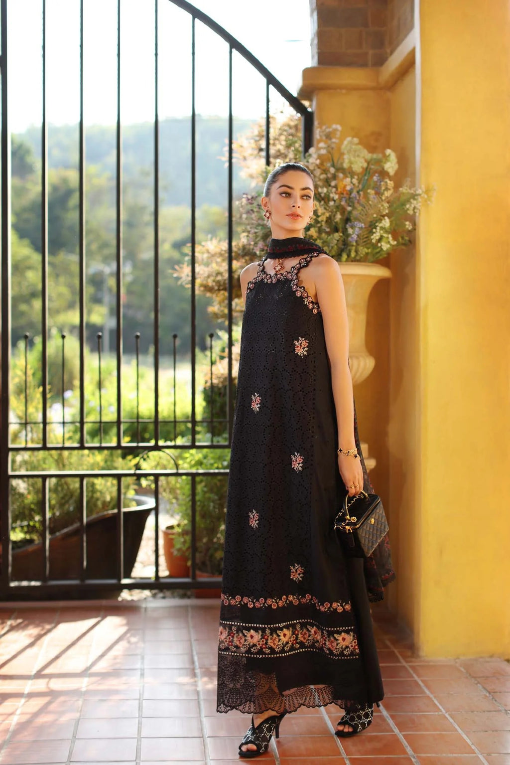 Woman in a black dress with floral embroidery standing in a sunlit room with a view of greenery outside.