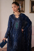 Woman in a dark blue embroidered outfit standing indoors with paintings on the wall.
