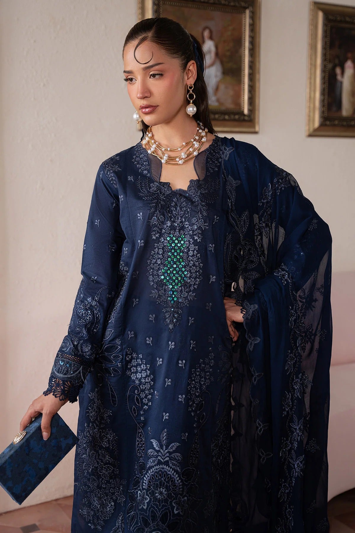 Woman in a dark blue embroidered outfit standing indoors with paintings on the wall.