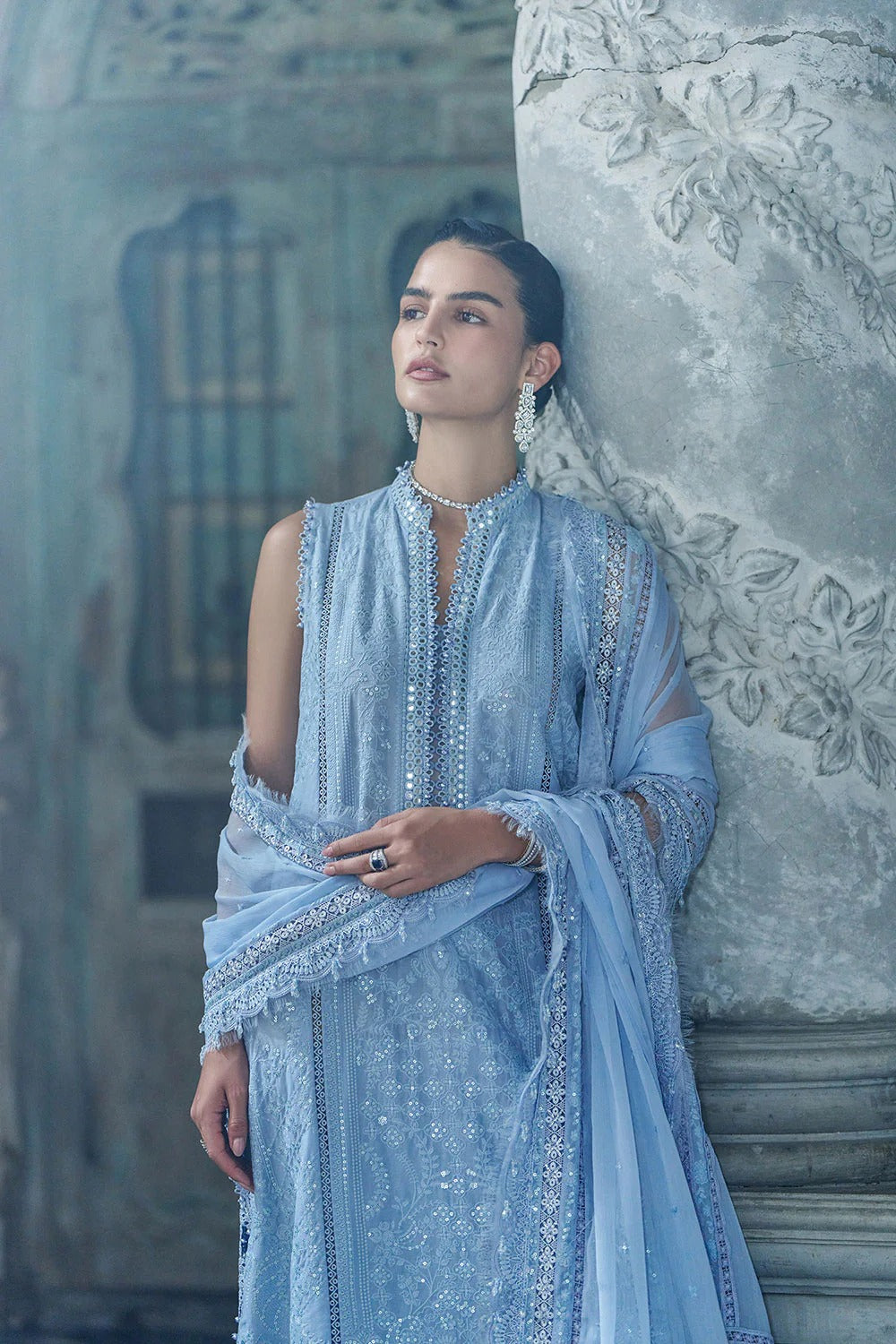 Woman in a light blue embroidered outfit leaning against an ornate wall.
