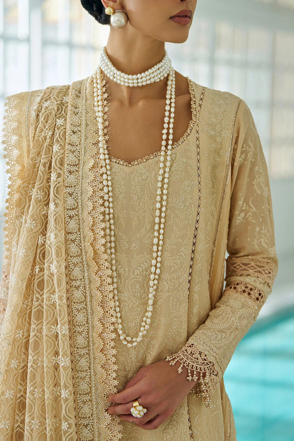 Woman wearing a beige embroidered outfit with a pearl necklace and earrings.