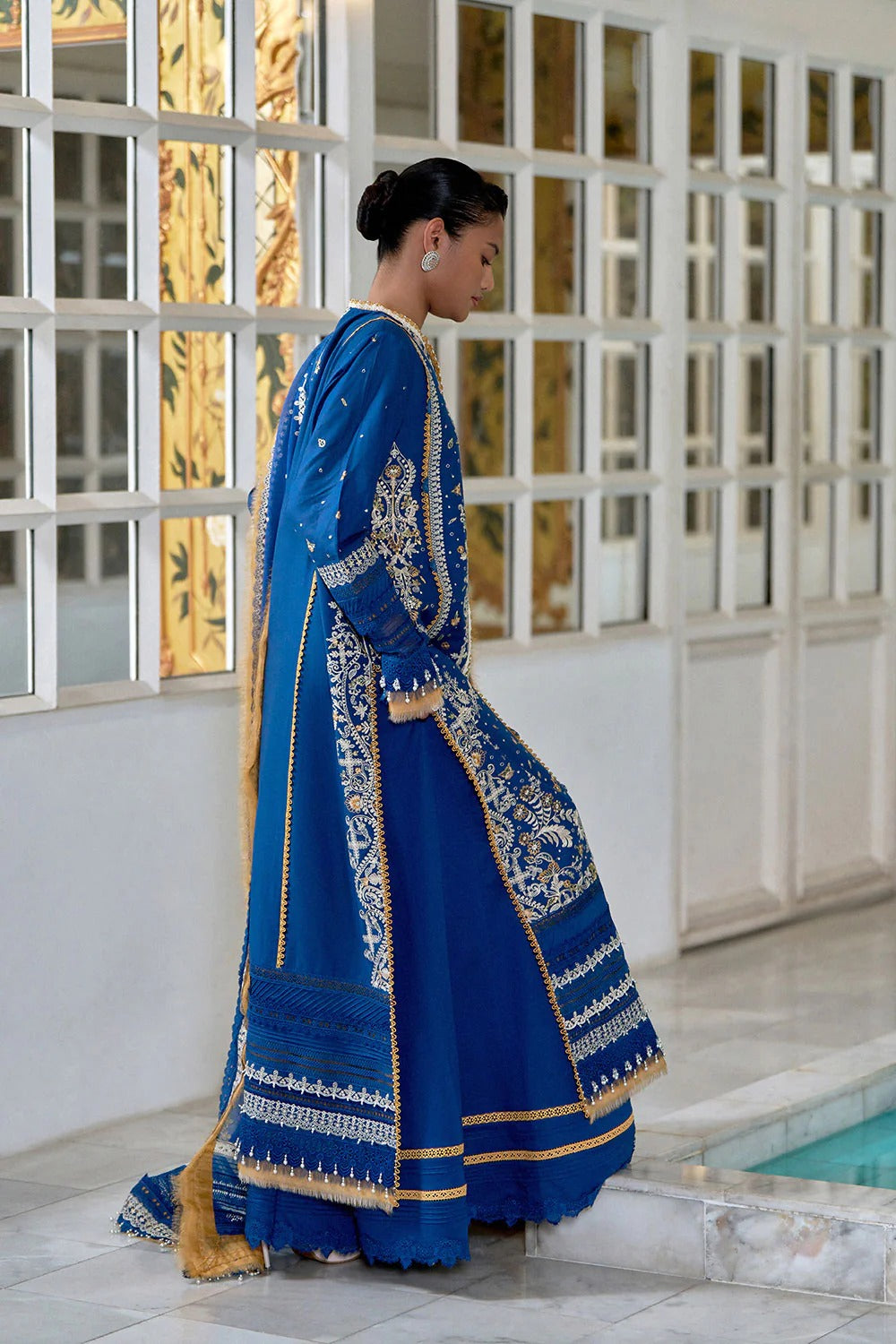 Woman in a blue traditional outfit with gold accents standing in front of a decorative wall.