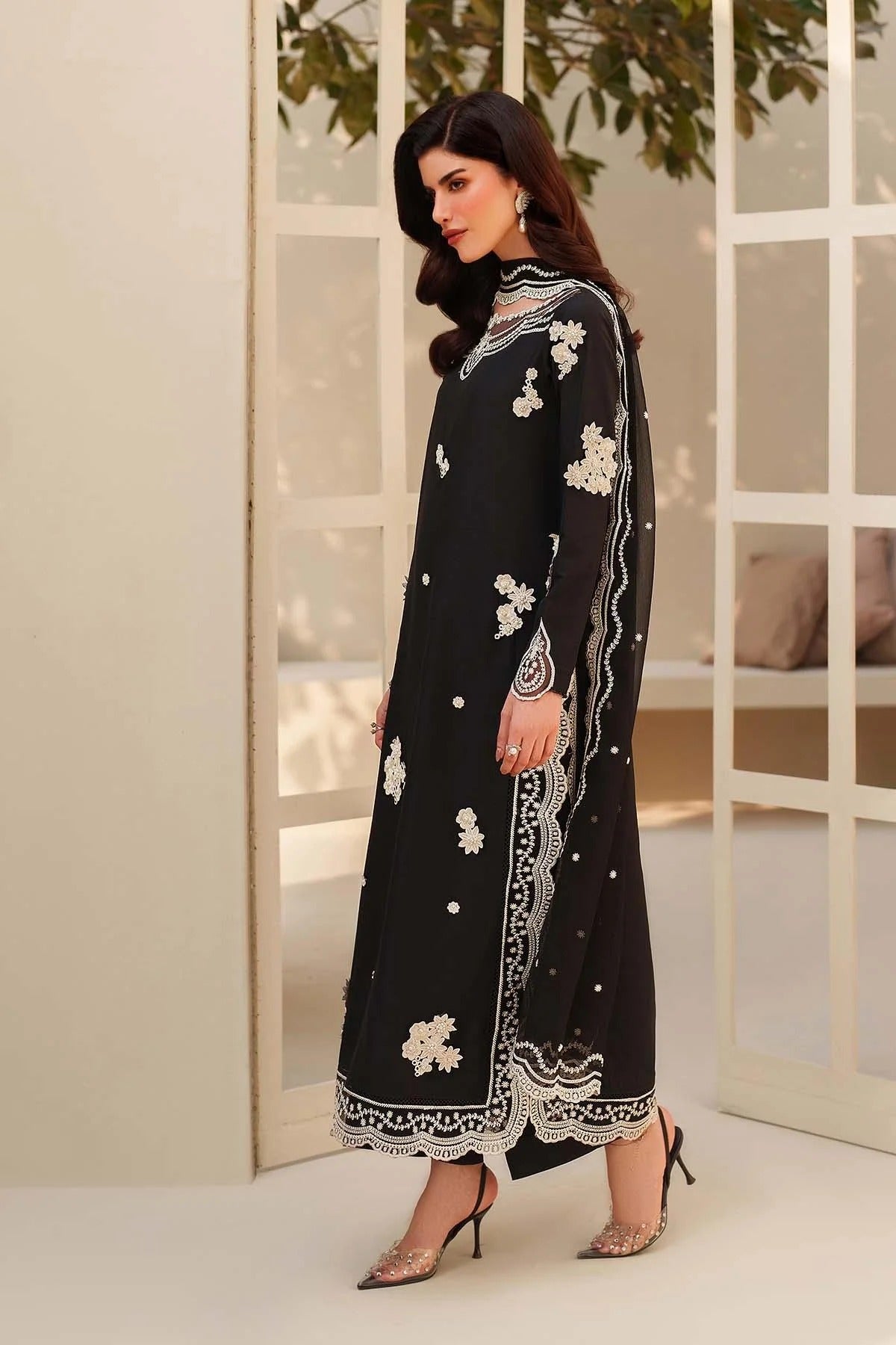Woman wearing a black embroidered outfit with white patterns in a minimal indoor setting.