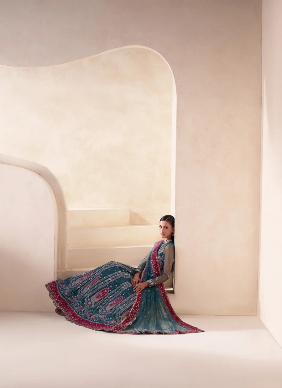 A woman sits on the steps of a minimalist cream-colored architectural structure, wearing a traditional South Asian outfit featuring a teal blue patterned skirt with a vibrant magenta border and a matching teal dupatta.