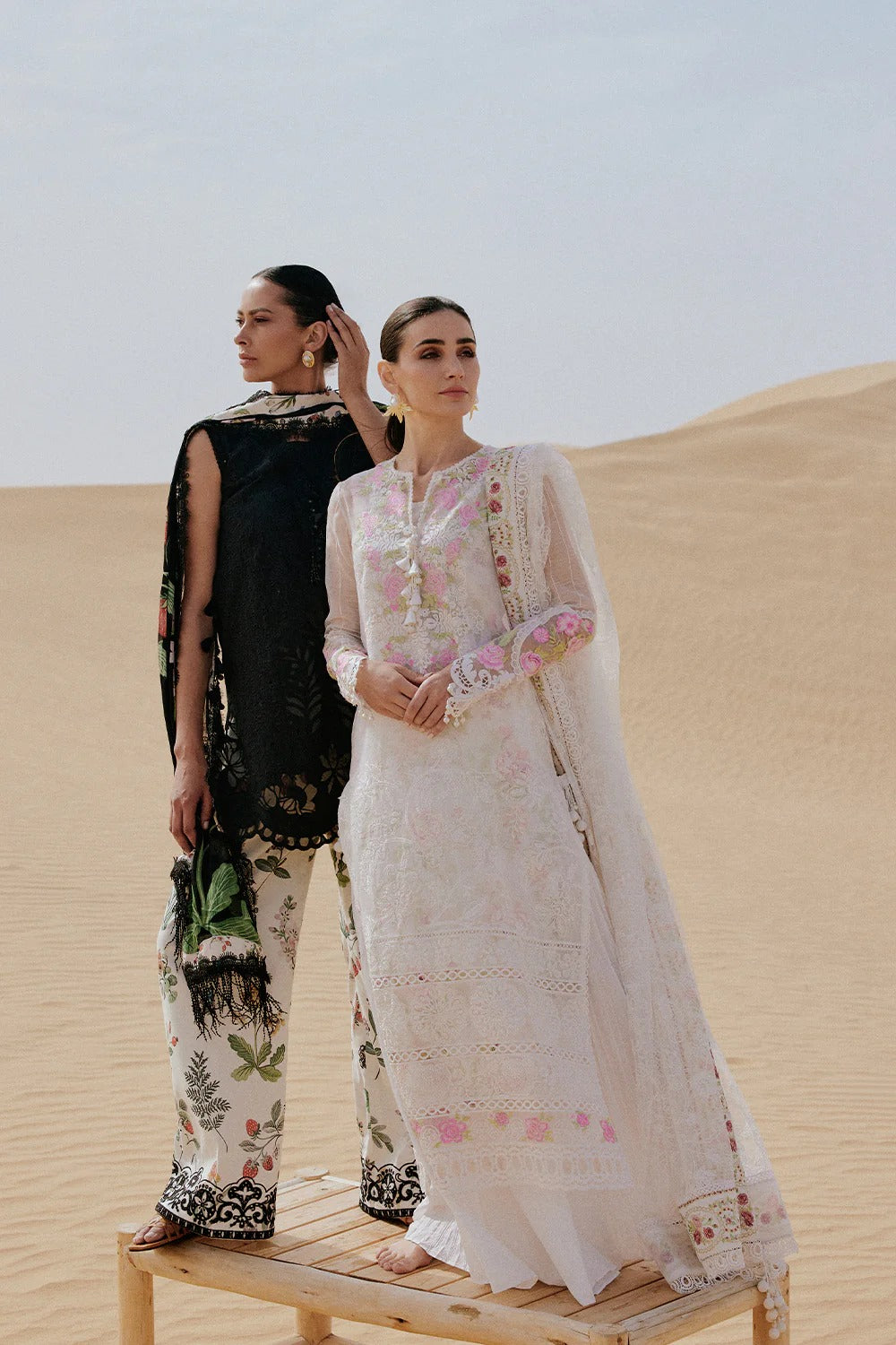 Two women in traditional outfits standing on sand dunes with a clear sky.