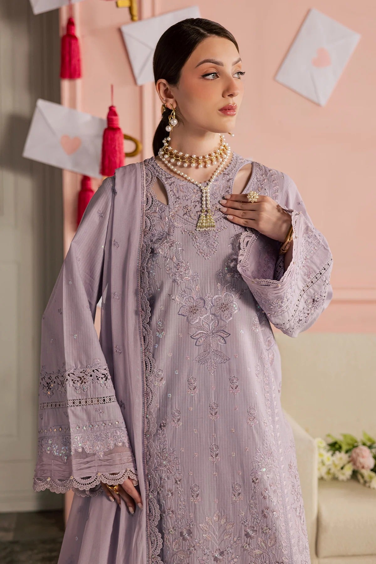 Woman in a gray embroidered outfit with a matching dupatta, holding a phone, against a pink wall.