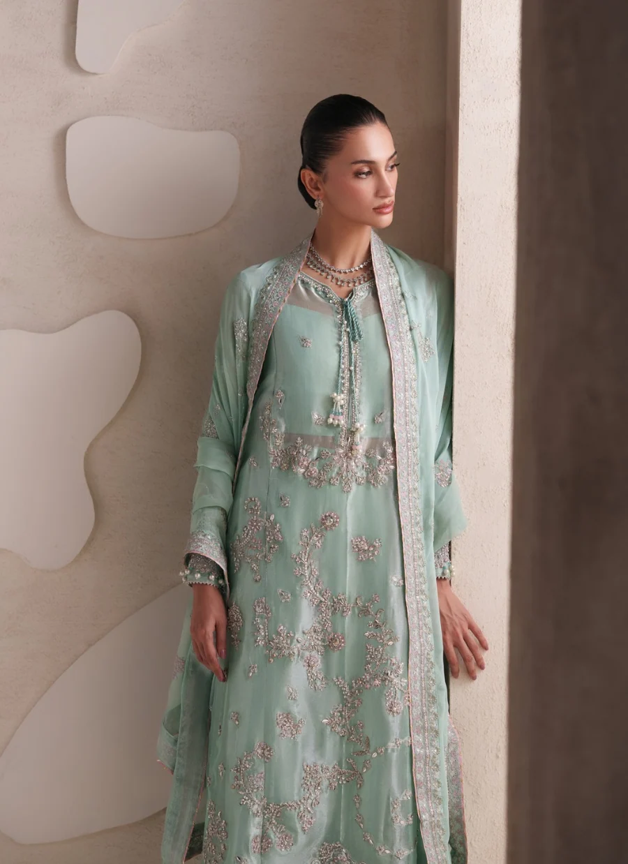 A woman models a mint green traditional outfit with intricate silver embroidery, featuring a long kurta, matching dupatta, and a light beige jacket draped over her shoulders, set against a textured beige wall with abstract shapes.