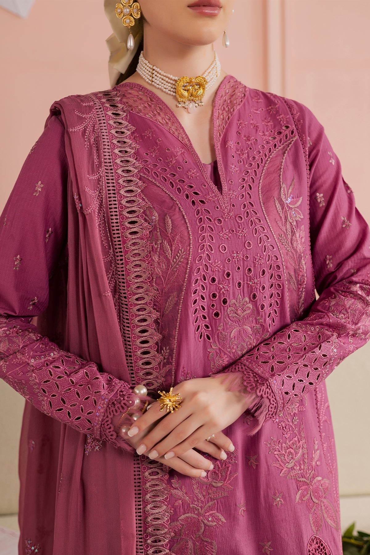 Close-up of a person wearing a pink embroidered traditional outfit with jewelry against a light pink background.