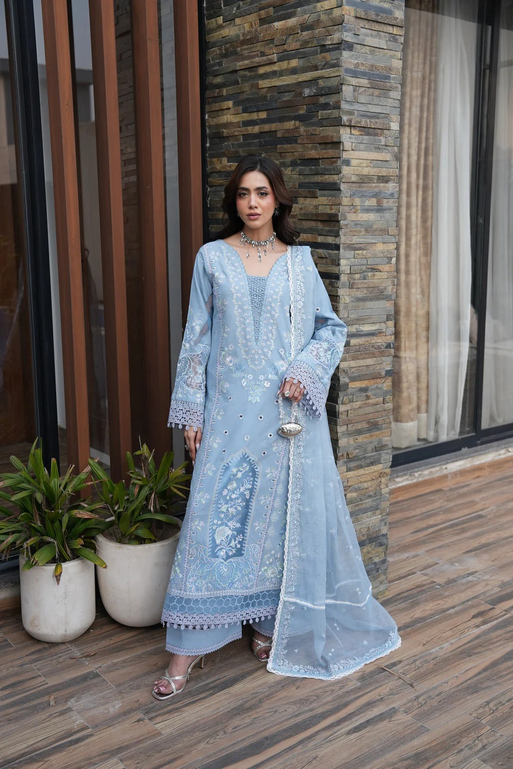 Woman in a light blue embroidered outfit standing in front of a textured wall.