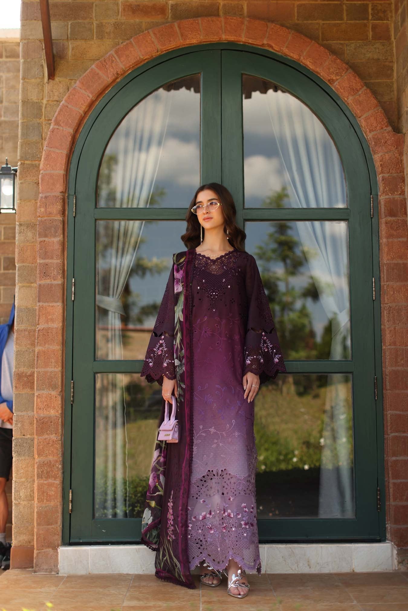 Woman in a purple dress standing in front of a large arched window with a brick building in the background.
