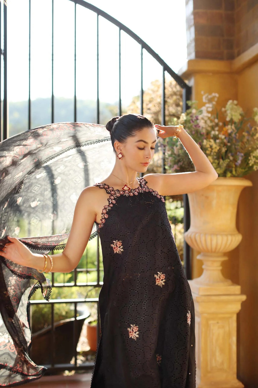 Woman in a black saree with floral patterns standing outdoors near a decorative column.