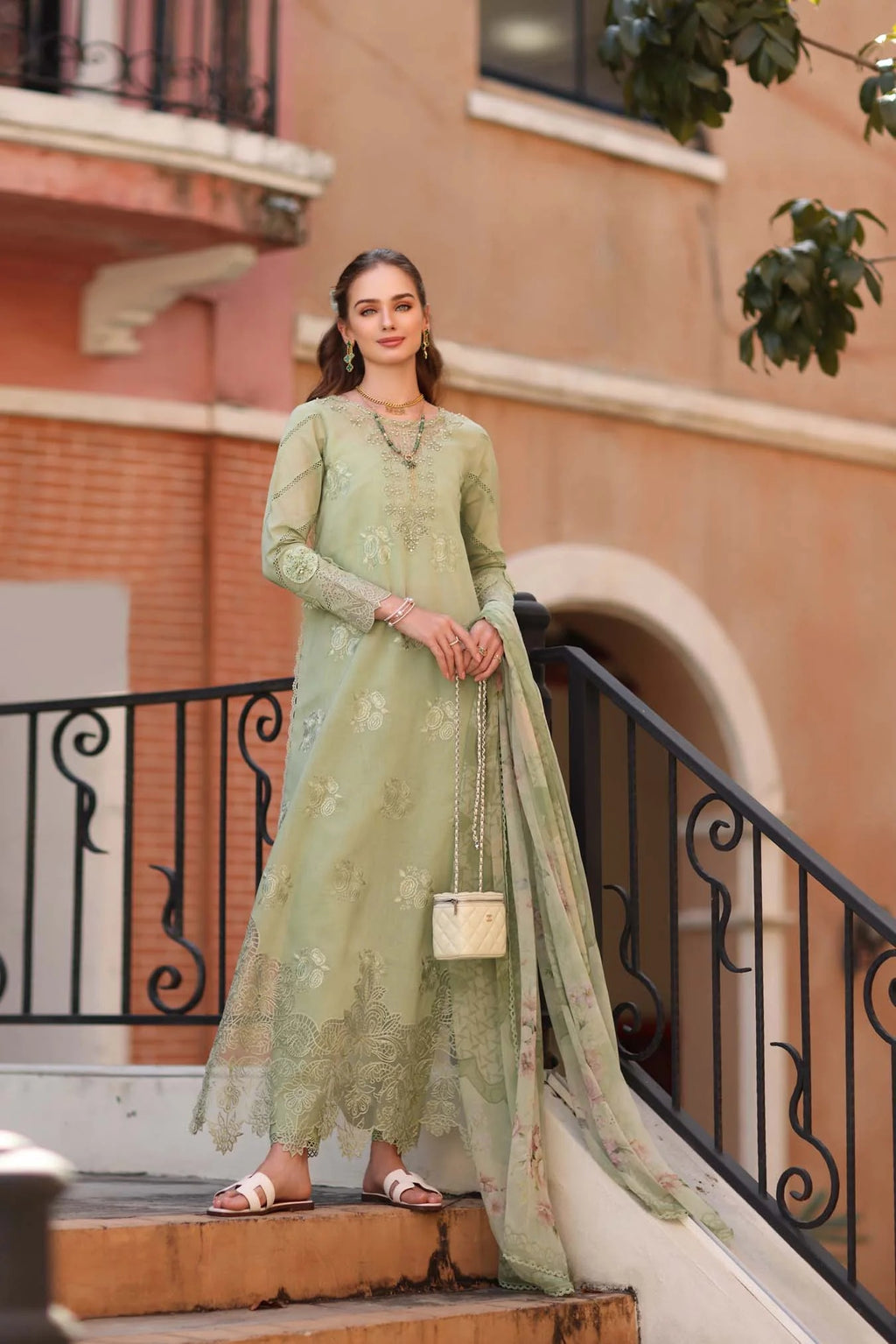 Woman in a light green traditional outfit standing on a balcony with a building in the background.