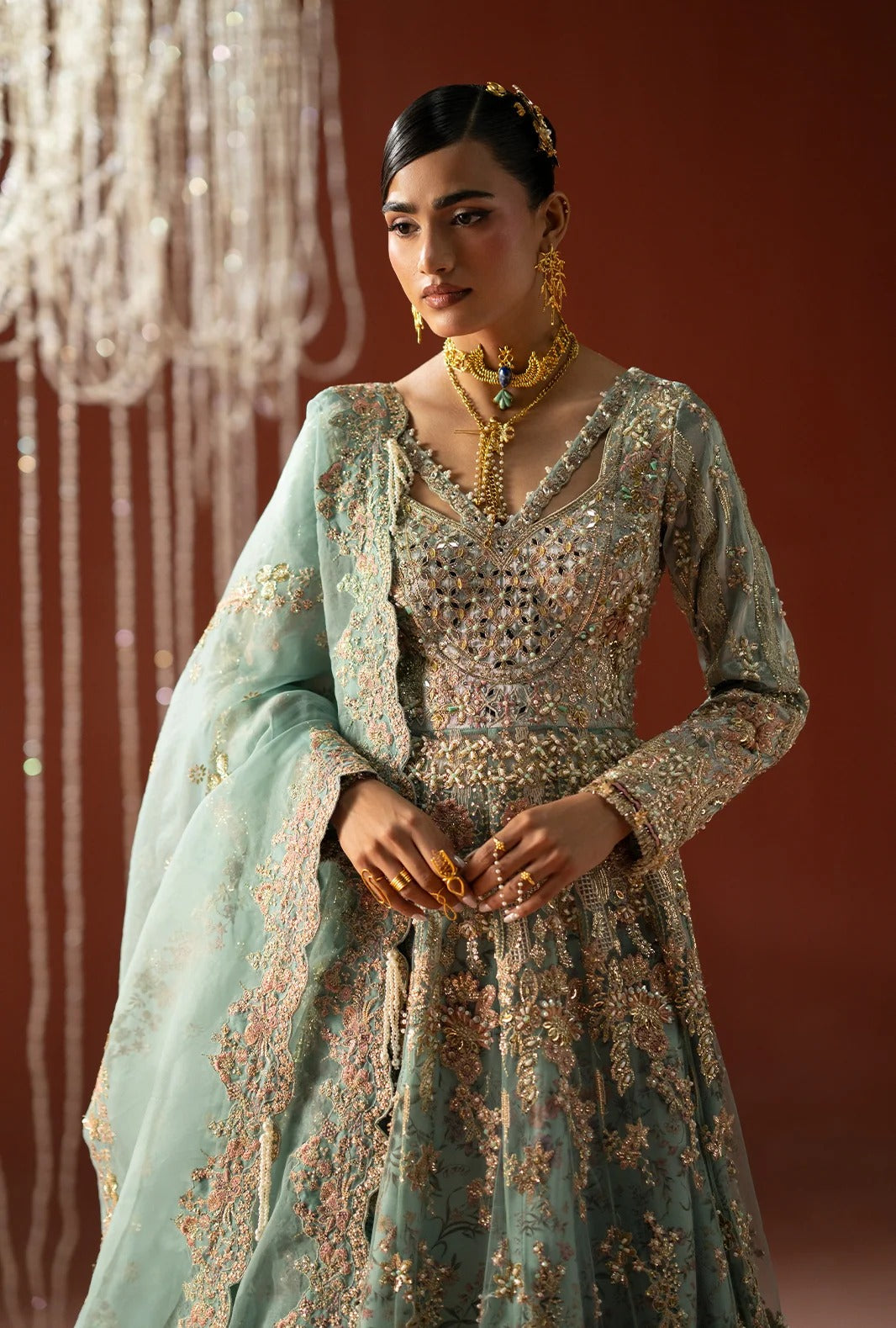 A woman models a traditional Indian anarkali gown in a dusty sage green color, heavily embellished with gold and silver embroidery. She is accessorized with a matching sheer dupatta, a gold necklace, and earrings, set against a warm brown background with hanging crystal decorations.