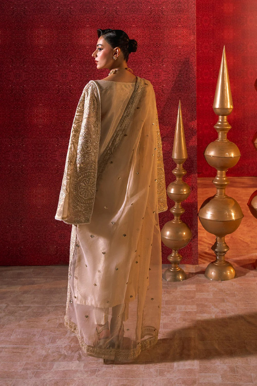 Woman in a traditional outfit standing against a red wall with decorative gold lamps.