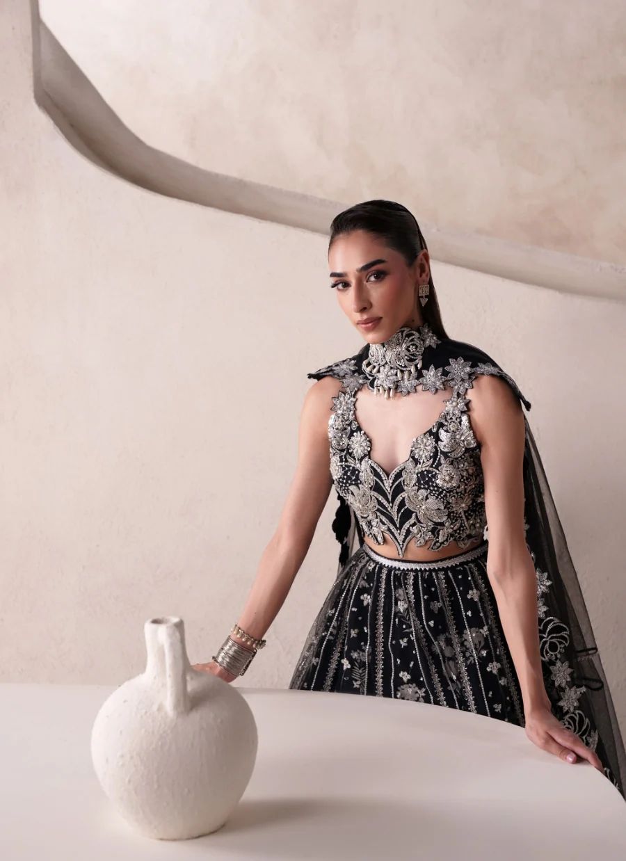 A woman models a black and silver heavily embellished traditional outfit with a sheer black dupatta, standing beside a white table holding a textured white vase against a beige architectural background.