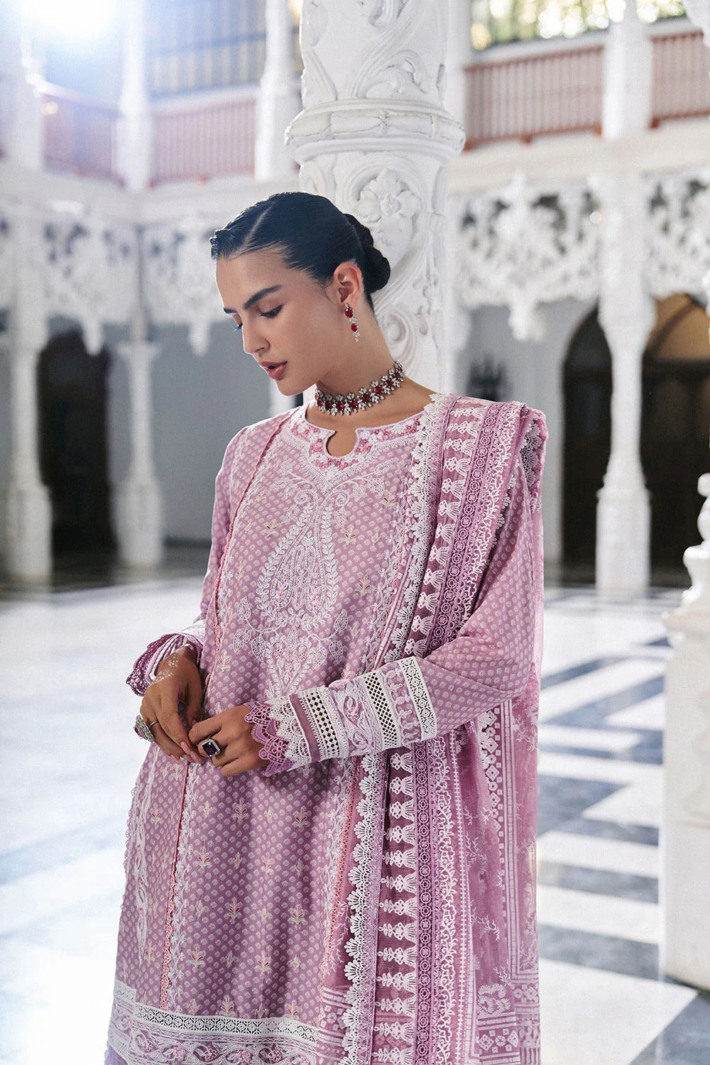 Woman in a pink traditional outfit with intricate patterns in an ornate indoor setting.