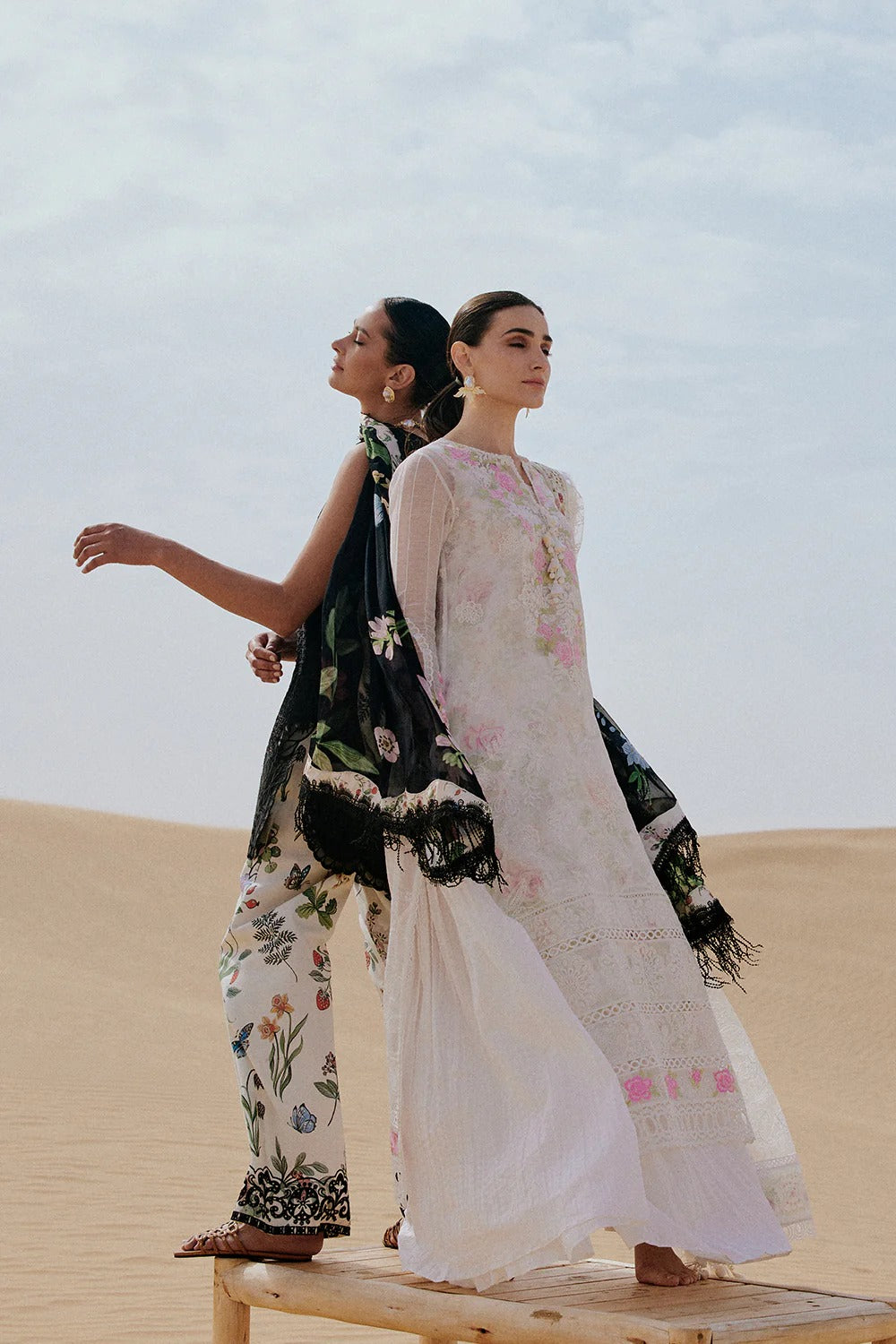 Two women in traditional outfits standing on sand dunes with a clear sky.