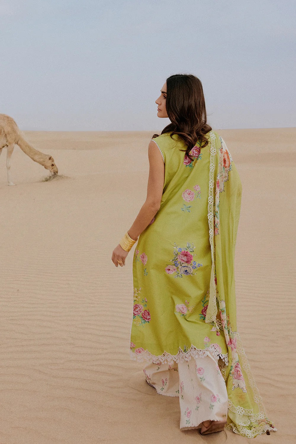 Woman in a green saree with floral patterns standing in a desert with a camel in the background.