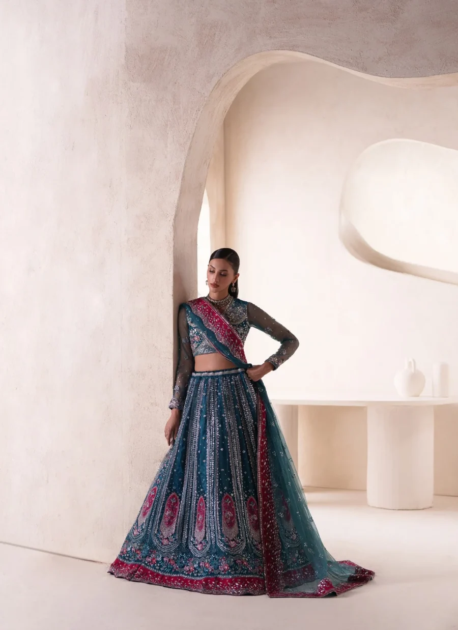 A woman models a traditional Indian lehenga choli set in a minimalist, warm-toned architectural setting. The outfit is a deep teal blue with intricate silver embroidery and vibrant magenta-pink borders, paired with sheer long sleeves.