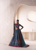 A woman models a traditional Indian lehenga choli set in a minimalist, warm-toned architectural setting. The outfit is a deep teal blue with intricate silver embroidery and vibrant magenta-pink borders, paired with sheer long sleeves.