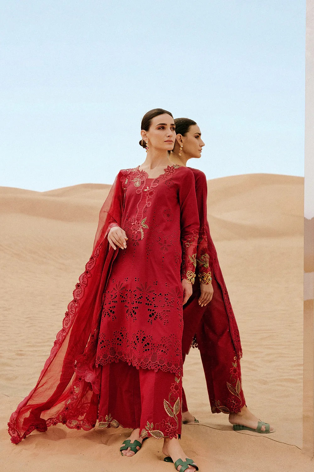 Two women in red traditional outfits standing in sand dunes with a clear blue sky.