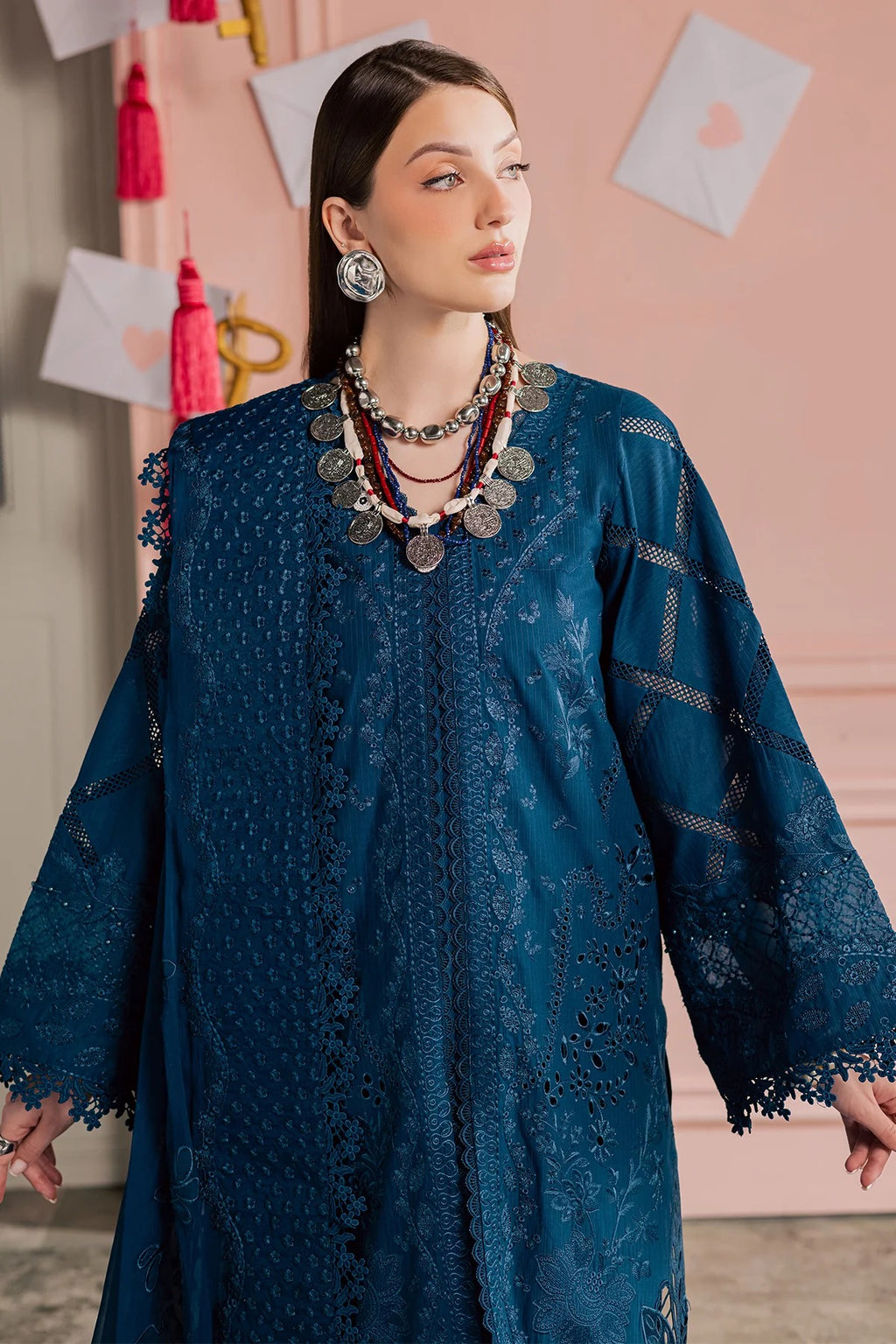 Woman wearing a blue embroidered dress with jewelry against a pink wall.