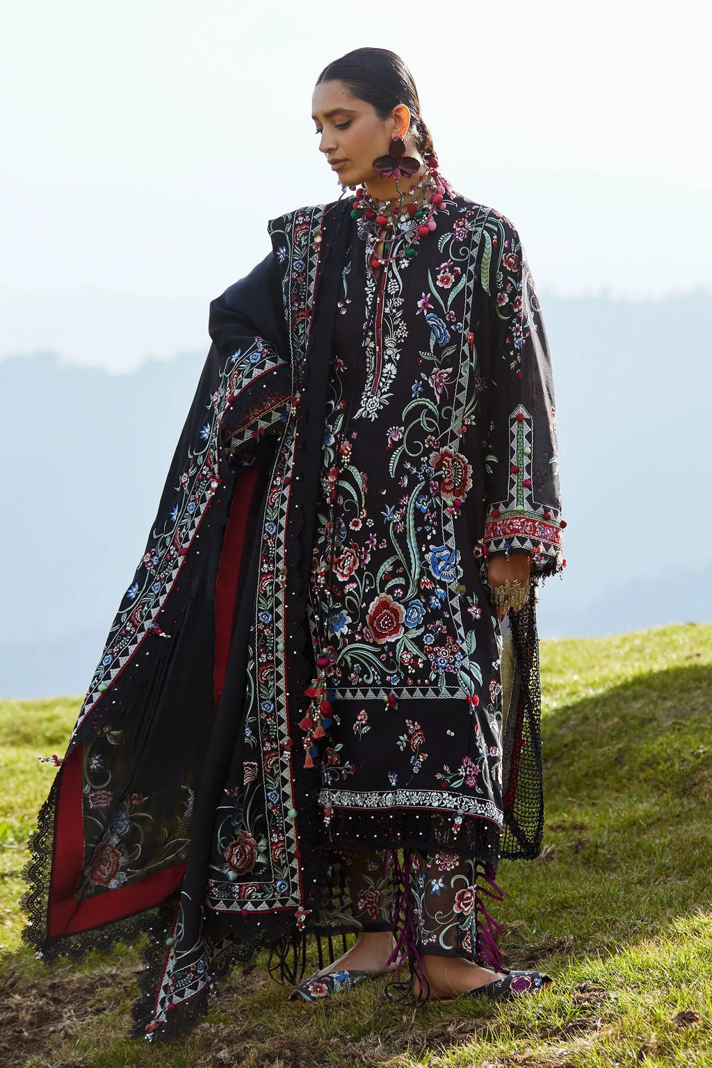 Woman in a traditional embroidered outfit standing on a grassy hill with mountains in the background