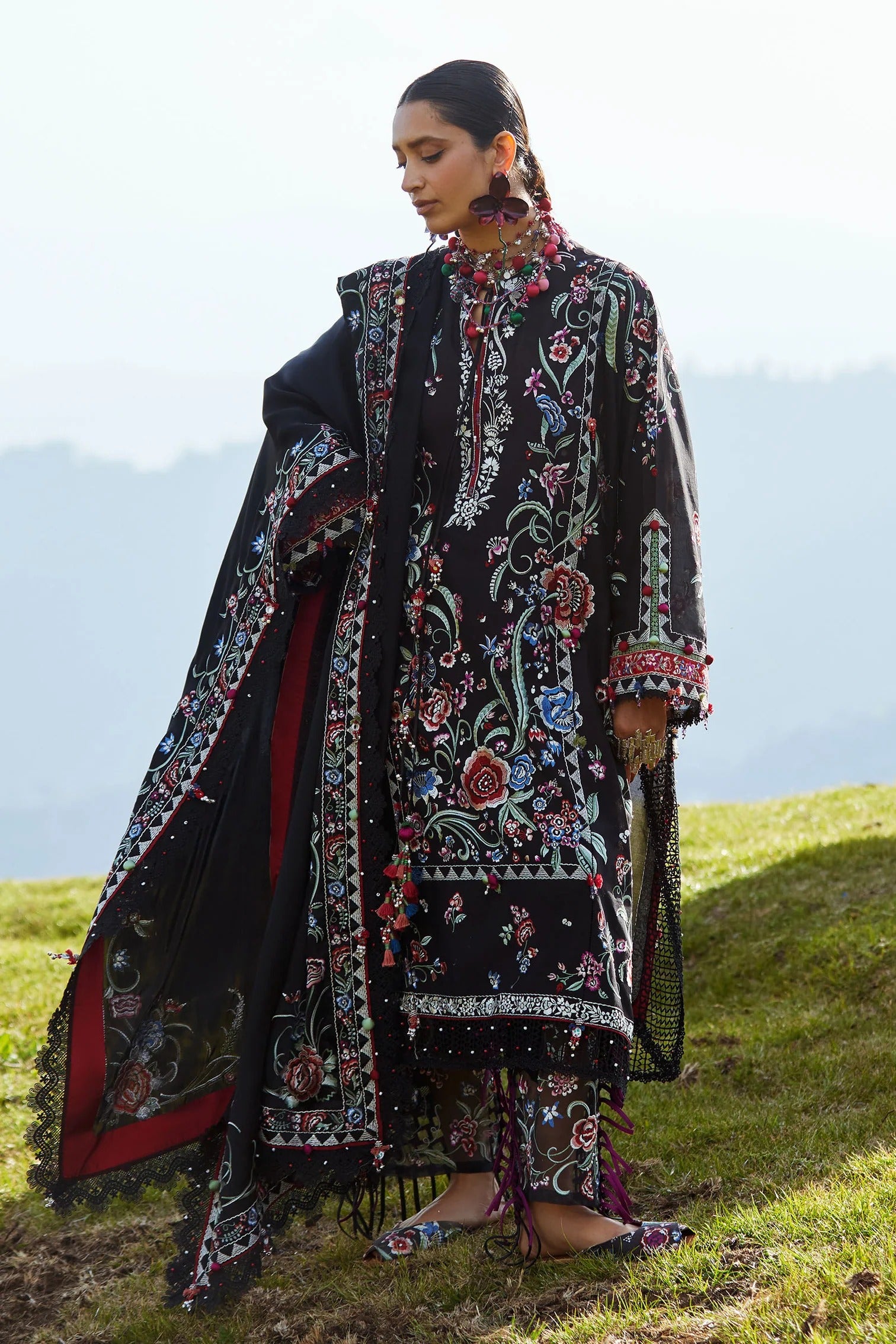 Woman in a traditional embroidered outfit standing on a grassy hill with mountains in the background