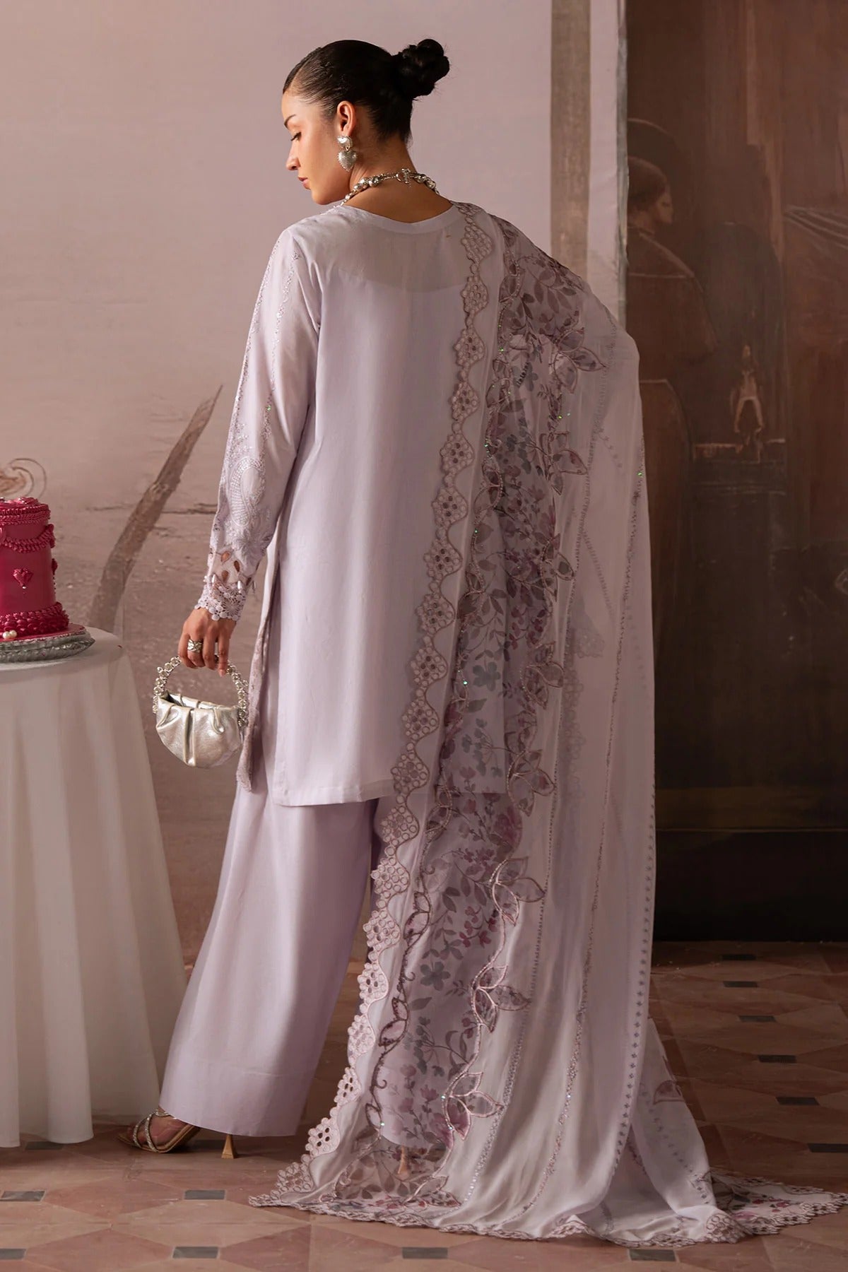 Woman in a white outfit with a floral shawl standing near a table with a red cake.
