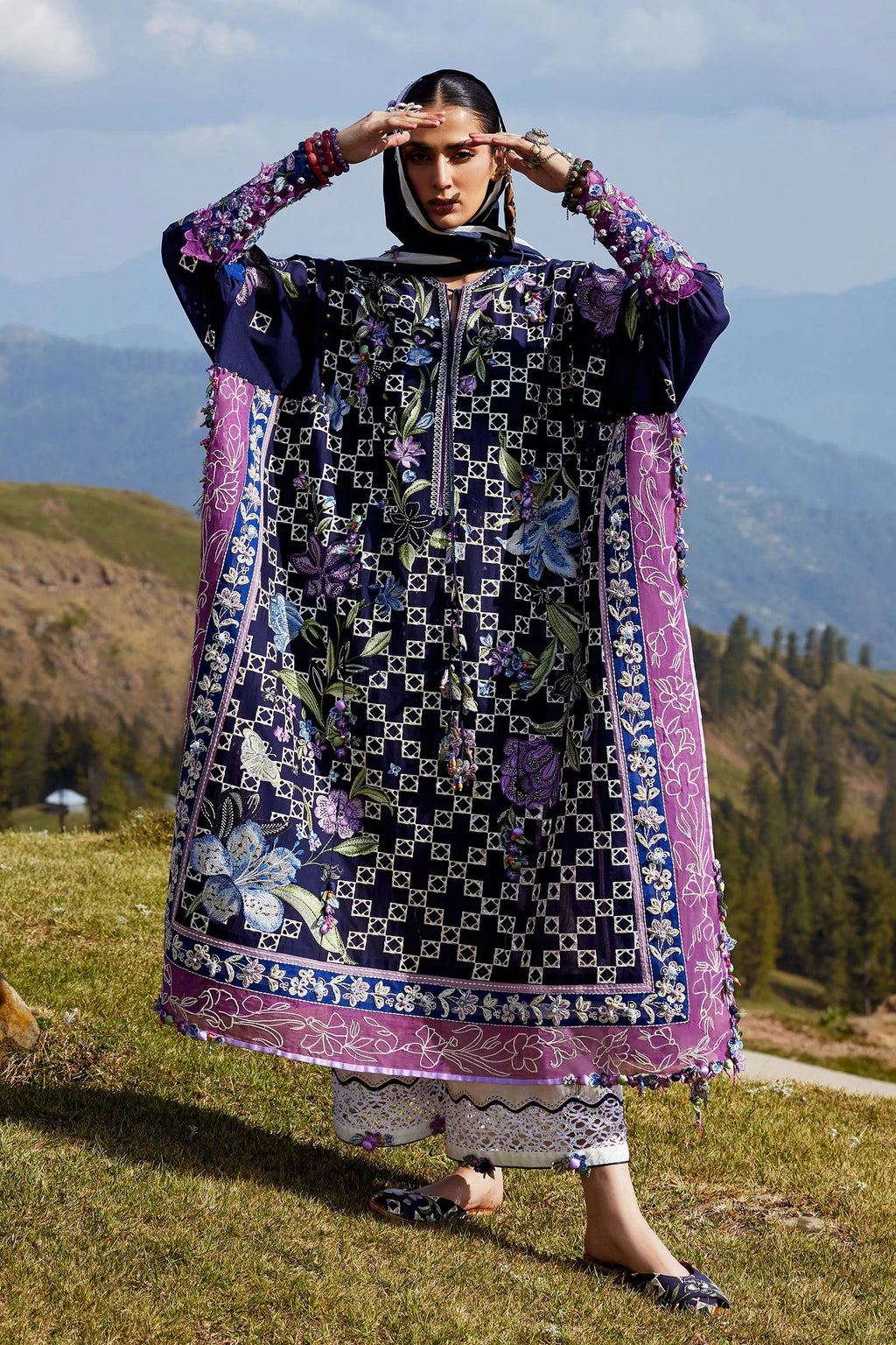 Woman in a traditional outfit with floral patterns standing in a scenic outdoor setting with mountains and greenery.