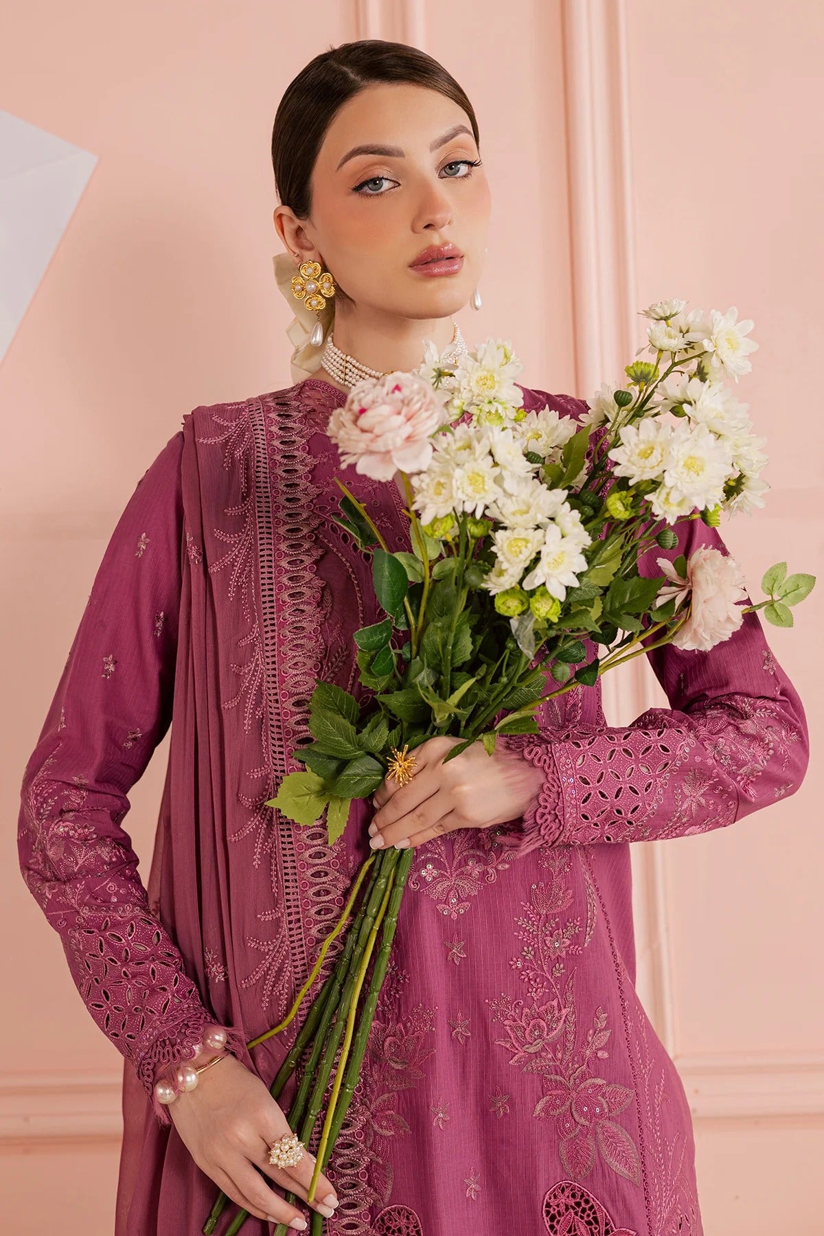 Woman in a purple embroidered outfit holding flowers against a pink background