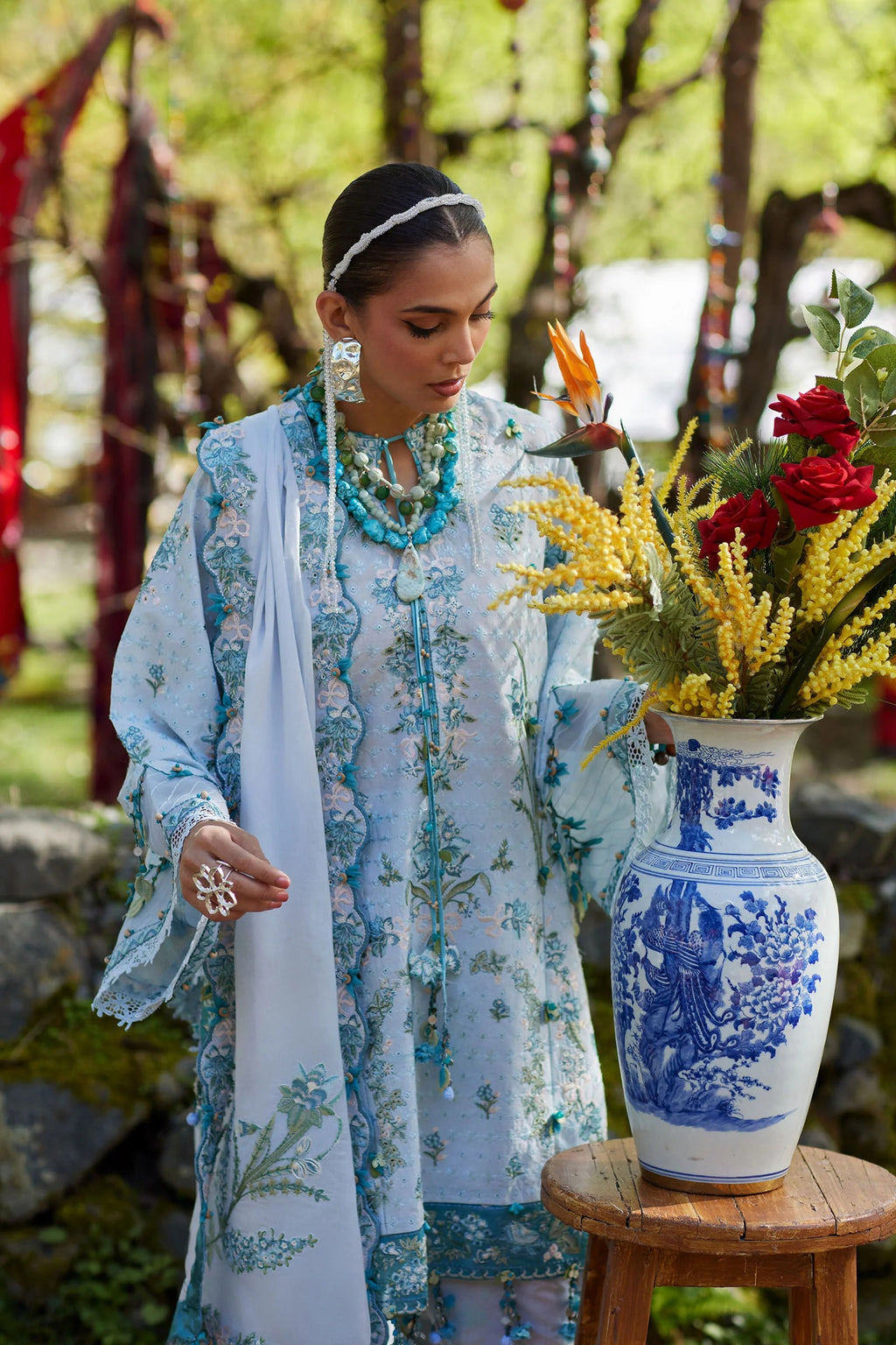 Woman in traditional attire holding flowers with a decorative vase in a natural setting