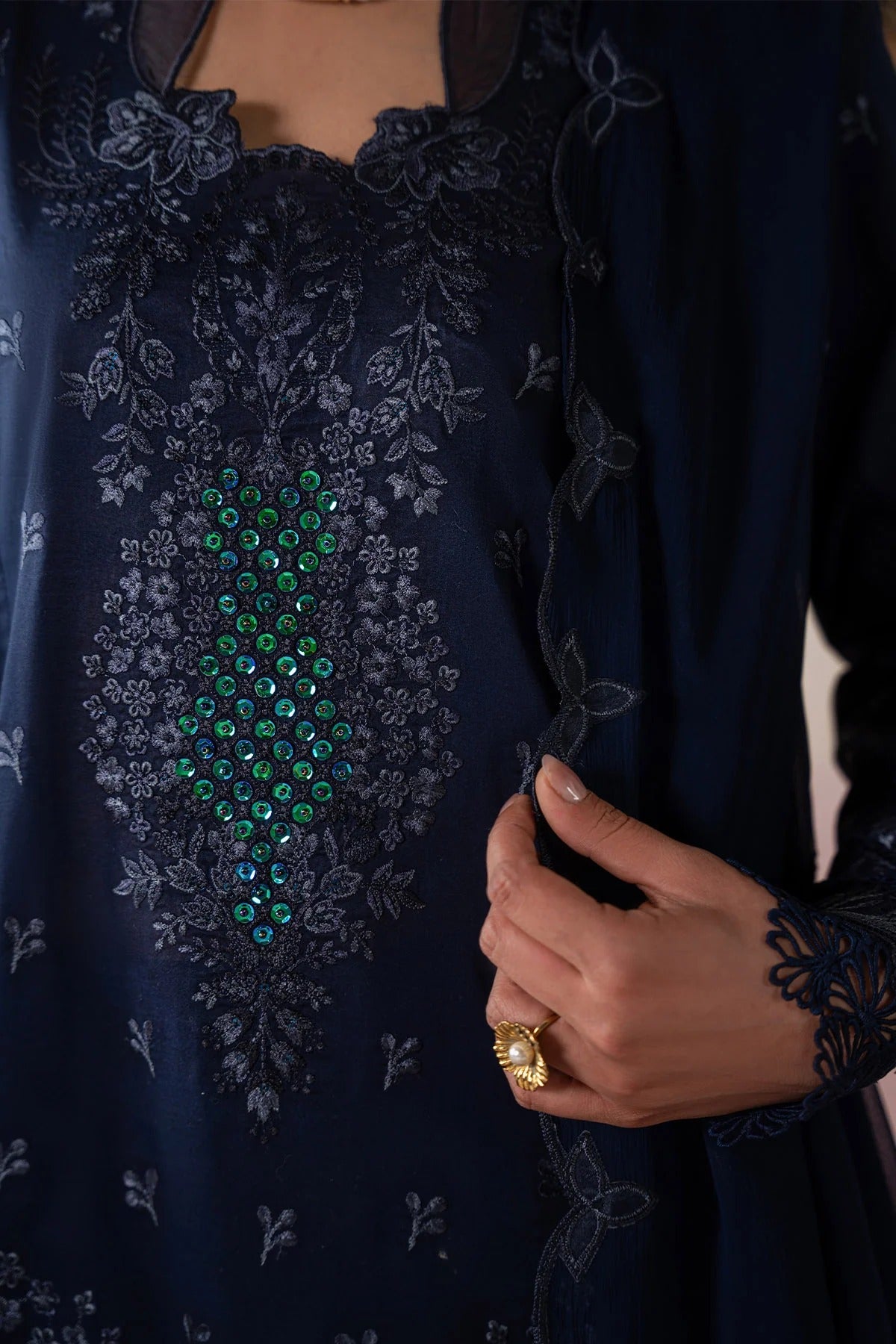 Navy blue embroidered garment with green gemstone details and a hand holding a phone.